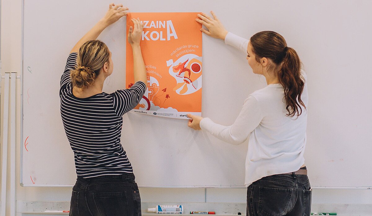 Divas sievietes pie tāfeles piestiprina projekta “Dizaina skola” plakātu.